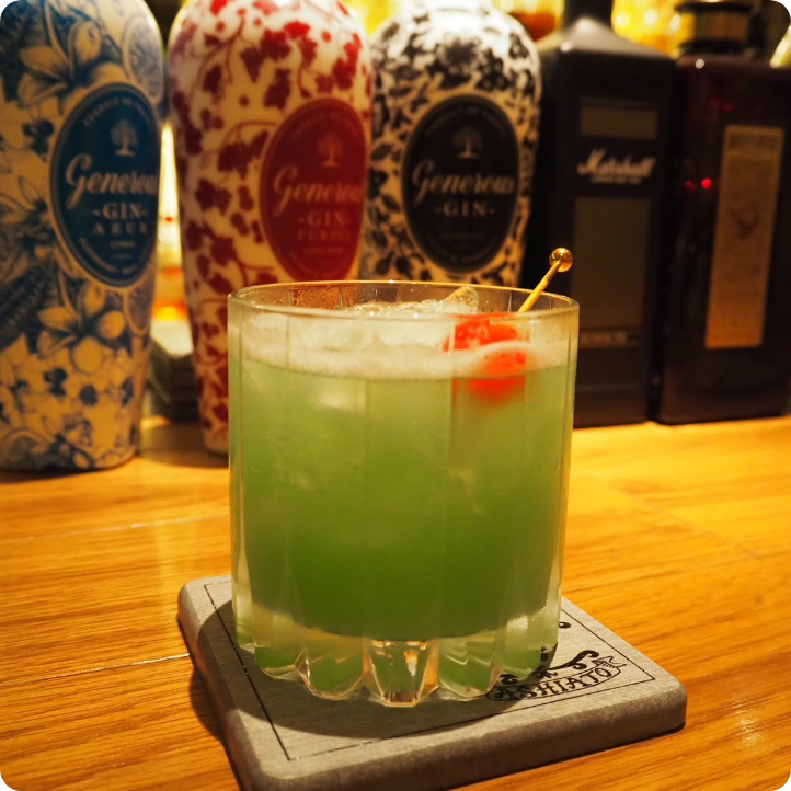 緑色のカクテルにチェリーを添えた一杯。華やかなジンボトルが並ぶバーカウンターでのドリンク